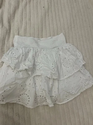 Vit volangkjol från Kappahl - Söt vit kortkjol från Kappahl med volanger och broderade detaljer. Kjolen har bred resår i midjan och är gjord i ett lätt bomullstyg med hålmönster som ger en somrig känsla.