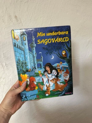 Min underbara sagovärld bok - Sagobok med hård pärm och färgglada illustrationer. Innehåller klassiska sagor som Törnrosa och Mästerkatten i stövlar. Omslaget är blått med motiv av prinsessa, djur och slott. Perfekt för dig som gillar magiska berättelser och fina bilder.