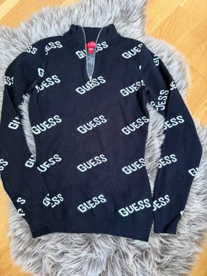 Svart stickad tröja från Guess - Säljer en svart stickad tröja från Guess med vit logotyp tryckt över hela tröjan. Tröjan har halv dragkedja med rund metallring. Storleken kan även passa till xs