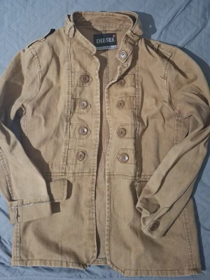 Beige militärinspirerad jacka Diesel - Snygg beige jacka från Diesel med militärinspirerad design, dubbla rader bruna knappar framtill och axelklaffar. Tillverkad i ett slitstarkt bomullstyg och har ståkrage för extra attityd. Perfekt för dig som vill sticka ut med en unik jacka.