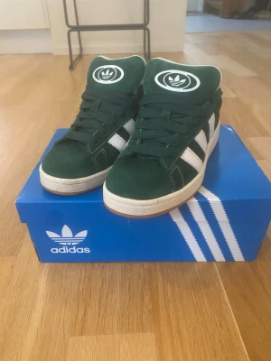 Adidas Campus gröna sneakers - Mörkgröna Adidas Campus sneakers i mocka med klassiska vita ränder på sidorna och vit sula. Skorna har gröna skosnören och Adidas-logga på plösen. Snygg retrostil som passar till det mesta och ger en clean look.