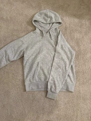 Ljusgrå basic hoodie med snören  - En stilren ljusgrå hoodie med huva och snören. Hoodien har en klassisk magficka framtill och ribbade muddar vid ärmar och nederkant. Tillverkad i mjukt sweatshirtmaterial.