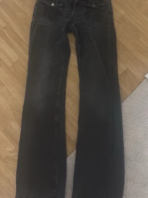 Svarta bootcut jeans med fickor - Säljer ett par svarta bootcut jeans med två framfickor med lock och silverfärgade knappar. Jeansen har en klassisk femficksmodell och är tillverkade i denim med lätt tvättad look. Perfekta för en avslappnad och trendig stil.