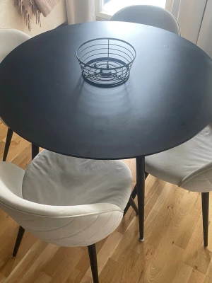 Runt matbord med fyra stolar - Säljer ett stilrent runt matbord med svart bordsskiva i metall och fyra matchande stolar med ljus klädsel och svarta ben. Perfekt för köket eller mindre matplats. Bordet har en modern design och stolarna är bekväma med mjuk sits. Pris kan diskuteras.