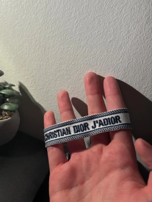 Dior armband - Snyggt armband från Christian Dior med texten 'J'ADIOR' i mörkblått på vit bakgrund. Armbandet är vävt i tyg och har dekorativa tofsar i ändarna. Perfekt accessoar för att lyfta din outfit med en exklusiv touch.