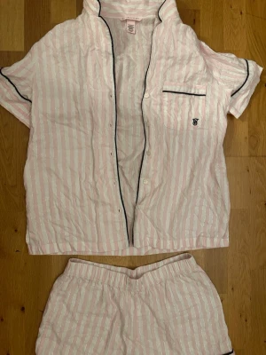 Randig pyjamas från Victoria's Secret - Säljer en söt pyjamas från Victoria's Secret i ljusrosa och vitt med silverränder. Setet består av en kortärmad skjorta med knappar och bröstficka samt matchande shorts. Svarta kontrastkanter ger en snygg touch. Perfekt för mysiga kvällar hemma.