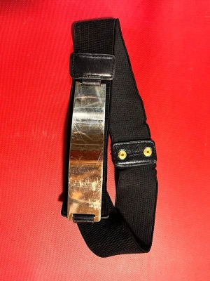 Svart elastiskt bälte med metallspänne - Säljer ett svart elastiskt bälte med brett band och ett stort, blankt metallspänne framtill. Bältet har tryckknappar i guld på baksidan för enkel stängning. Perfekt accessoar för att ge outfiten en cool touch.