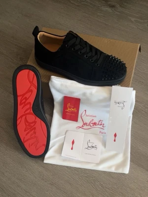  Christian louboutin skor  - Snygga svarta sneakers från Christian Louboutin med klassisk röd sula och coola svarta nitar på tån. Skorna har snörning, låg profil och är tillverkade i mocka med läderdetaljer. Perfekta för dig som vill sticka ut med en edgy och exklusiv look.