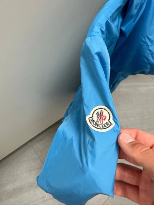 Blå vindjacka från Moncler Storlek 14y - Snygg blå vindjacka från Moncler med huva och dragkedja framtill. Jackan har två sidofickor med knappar och elastiska muddar vid ärmar och midja. Tillverkad i lätt material och har Moncler-logga på ärmen. Perfekt för vår,sommar och för hösten som kommer!