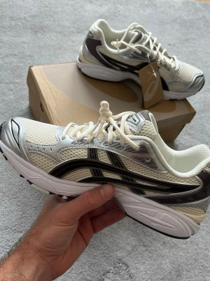Asics Gel-Kayano 14 Cream/Black - Snygga sneakers från Asics i vitt och silver med svarta detaljer. Ovandelen är i mesh och syntetmaterial för bra andningsförmåga och komfort. Klassisk låg modell med tydliga Asics-loggor på sidorna och en rejäl, stötdämpande sula.