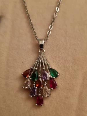 Halsband med färgglada droppformade stenar - Snyggt halsband i silverfärgad metall med ett hänge dekorerat med flera droppformade stenar i olika färger som rött, grönt, lila, orange och klart. Hänget har en unik design där stenarna hänger i små grupper från tunna kedjor. Perfekt för dig som gillar färg och vill sticka ut.