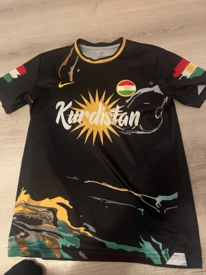 Svart Kurdistan t-shirt med tryck - Snygg svart t-shirt med Kurdistan-tryck och solsymbol på bröstet. Färgglada detaljer i gult, grönt, rött och vitt samt Kurdistans flagga på ärmarna. Tillverkad i ett mjukt och stretchigt material, perfekt för sport eller träning.
