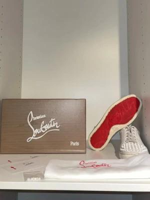 Vita sneakers med nitar från Louboutin - Säljer ett par vita sneakers från Christian Louboutin med ikoniska röda sulor och coola nitar över hela tån. Skorna är i läder och har klassisk låg modell med snörning. Perfekta för dig som vill sticka ut med en edgy och lyxig stil. Dom är äkta. Pris går att diskutera.
