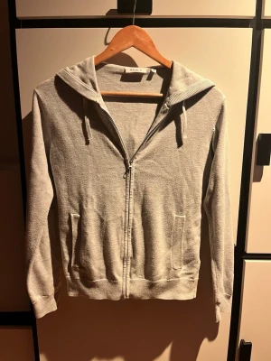 Ljusgrå zip hoodie med huva från RYVLS - En stilren ljusgrå hoodie med dragkedja framtill och två fickor. Hoodien har en klassisk huva med dragsko och ribbade muddar vid ärmslut och nederkant. Perfekt för en avslappnad look och enkel att matcha med det mesta.