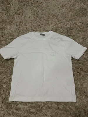 Vit basic t-shirt med diskret logga - Säljer en stilren vit t-shirt i bomull med korta ärmar och rund hals. På bröstet finns en liten, diskret broderad logga i vitt. Perfekt för dig som gillar minimalistisk och clean stil.