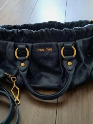 Svart handväska från Miu Miu - Snygg svart handväska från Miu Miu i glansigt läder med guldfärgade detaljer och dubbla handtag. Väskan har en rynkad ovankant och avtagbar axelrem. Perfekt för dig som vill ha en lyxig och trendig accessoar.