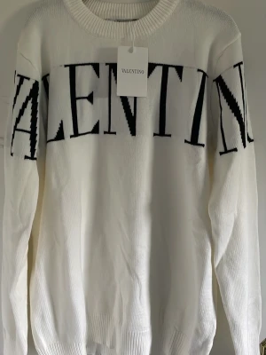 Vit stickad tröja från Valentino - Säljer en vit stickad tröja från Valentino med stor svart logotyp över bröstet. Tröjan har rund halsringning och ribbade muddar vid ärmslut och nederkant. Tillverkad i Italien och har en klassisk, avslappnad passform. Passar perfekt nu till hösten. Storleken passar L och M