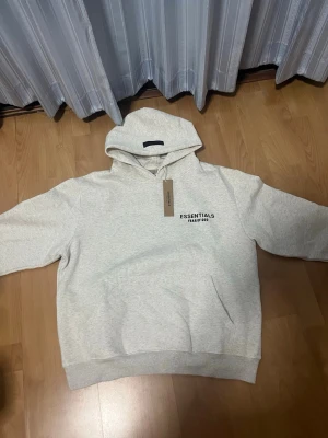 Ljusgrå hoodie Essentials Fear of God - Snygg ljusgrå hoodie från Essentials Fear of God med svart logga tryckt på bröstet. Klassisk modell med huva och ribbade muddar vid ärmslut och nederkant. Tillverkad i mjukt material för maximal komfort. Perfekt för en avslappnad och trendig look.