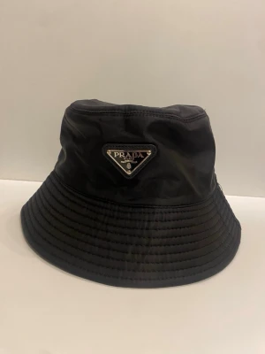 Svart bucket hat från Prada - Snygg svart bucket hat från Prada i glansigt nylonmaterial. Klassisk form med bred skärm och ikoniska metalltriangeln med logga framtill. Perfekt accessoar för en trendig och avslappnad stil.