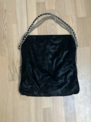 Svart handväska med kedjedetaljer - Snygg svart handväska i mjukt material med silverfärgade kedjor som handtag och längs kanterna. Väskan har en fyrkantig form och ger en cool och trendig vibe. Perfekt för dig som gillar statement-accessoarer och vill ha plats för allt du behöver.