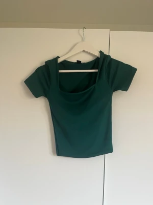 Mörkgrön croppad topp med fyrkantig ringning - Snygg mörkgrön croppad topp med korta ärmar och fyrkantig ringning. Perfekt till jeans eller kjol för en trendig look. Materialet känns mjukt och stretchigt, vilket gör den bekväm att bära.