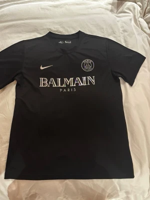 Svart PSG x Balmain t-shirt - Snygg svart t-shirt med PSG och Balmain Paris-logga på bröstet samt Nike swoosh. Klassisk rund halsringning och korta ärmar. Perfekt för dig som gillar streetwear och sportmode.