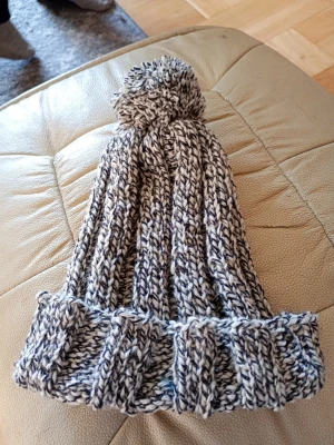 Gråmelerad stickad mössa med tofs - Säljer en ribbstickad mössa i gråmelerat garn med stor tofs på toppen. Mössan har bred uppvikt kant och är gjord i tjockt, mjukt material som håller dig varm. Perfekt för kalla dagar och ger en avslappnad, trendig look. Ull polyester raggsocksgarn. Hemstickad.