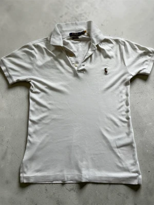 Vit pikétröja från Polo Ralph Lauren - Klassisk vit pikétröja från Polo Ralph Lauren i slim fit-modell. Tröjan har korta ärmar, krage och knappar framtill samt den ikoniska broderade loggan på bröstet. Tillverkad i mjuk bomull som känns skön mot huden. Den har två små hål som man kan se på bilden men verkligen inget som syns när man har på sig tröjan👍 priset är ej hugget i sten så kom med bud🤝
