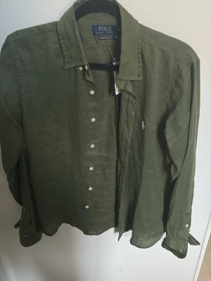 Olivgrön linneskjorta Polo Ralph Lauren - Snygg olivgrön skjorta från Polo Ralph Lauren i linne. Klassisk krage, vita knappar framtill och lång ärm med knapp vid manschetten. RL Untucked Fit för en avslappnad look. Liten broderad logga på bröstet. Helt ny, aldrig använt. Köpt i Usa när min syster var där och den passar ej därav säljer jag den.