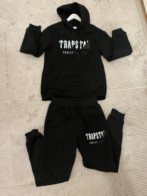 Svart loungewear set med tryck - Säljer ett svart loungewear set med hoodie och byxor. Hoodien har huva, magficka och vitt tryck med texten 'TRAPSTAR' på bröstet. Byxorna har resår i midjan, dragsko och matchande tryck på benet. 