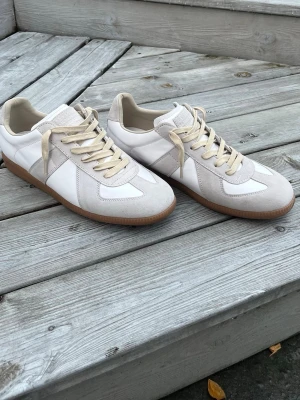Maison margiela skor  - Snygga sneakers i vitt och beige från Maison Margiela. Ovandelen är i en mix av mocka och läder med beige detaljer och vita paneler. Skorna har beige snören och en klassisk gummisula. Perfekta för en clean och stilren look.