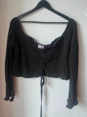 Svart croppad ribbad off shoulder topp - Snygg svart croppad off shoulder topp med långa ärmar och ribbad struktur. Toppen har snörning framtill och vågig kant nertill samt vid ärmsluten. Perfekt för dig som gillar trendiga detaljer och en figurnära passform.