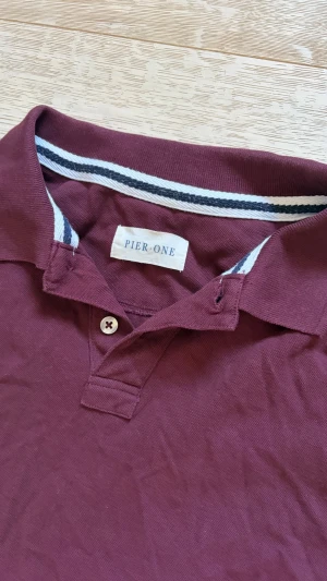 Vinröd pikétröja från Pier One - Snygg vinröd pikétröja från Pier One med klassisk krage och knappslå. Tröjan har korta ärmar och är tillverkad i mjuk bomull. Perfekt för en avslappnad men stilren look.