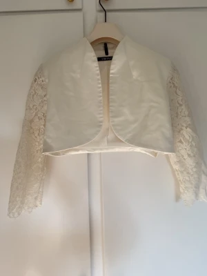 Vit bolero med spetsärmar från Swing - Kort vit bolerojacka från Swing med glansigt tyg framtill och långa spetsärmar. Jackan har öppen front och elegant, rundad krage. Perfekt för att lyfta en outfit med en lyxig touch.