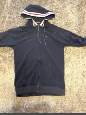 Mörkblå hoodie med dragkedja och huva - Snygg mörkblå hoodie med dragkedja och fickor framtill. Huvan har dragsko och ett vitt, rött och blått band längs kanten. Perfekt för en avslappnad stil. Materialet ser ut att vara mjukt och bekvämt, troligen bomullsblandning.