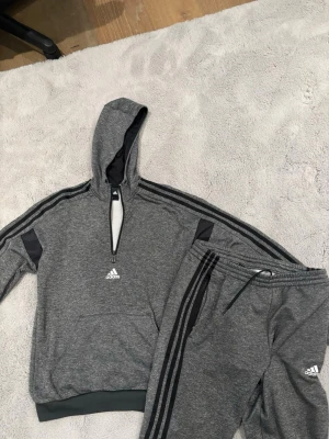 Adidas tracksuit - Säljer ett grått loungewear set från Adidas med matchande hoodie och byxor. Setet har svarta detaljer och klassiska Adidas-ränder längs ärmar och ben samt logga på både tröja och byxa. Perfekt för chill dagar hemma eller när du vill vara bekväm.