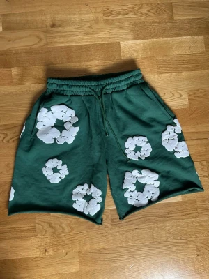 Gröna shorts med vita blommor - Säljer ett par gröna shorts med stora vita blommönster. Shortsen har resår i midjan och snörning framtill för skön passform. Perfekta för varma dagar och ger en chill vibe till din outfit.