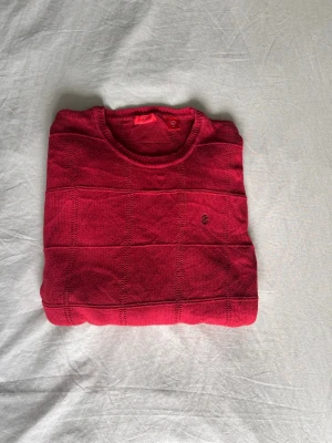 IZOD Röd crewneck-jumper - Snygg röd stickad Izodtröja med rund hals och diskret rutmönster. Tröjan har långa ärmar och ribbade muddar vid ärmslut och nederkant. Liten broderad detalj på bröstet ger en stilren touch. Perfekt för dig som vill ha en enkel men trendig look.