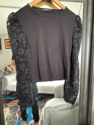 Svart topp med spetsärmar - Snygg svart topp med långa, transparenta spetsärmar i blommigt mönster. Kroppen är i mjuk bomull och toppen har en rak, croppad passform. Perfekt för dig som vill ha en trendig och unik look.