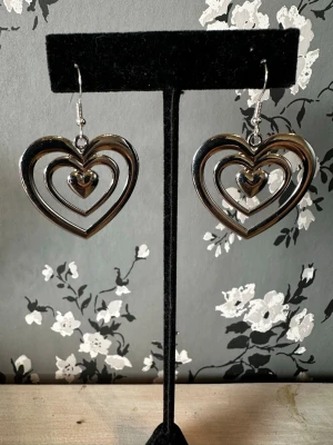 Silvriga hjärtformade örhängen - Säljer ett par coola örhängen i silverfärgad metall med dubbla hjärtan och en liten hängande hjärtberlock i mitten. De har en modern och trendig look med öppen design och passar perfekt för dig som gillar statement-smycken.