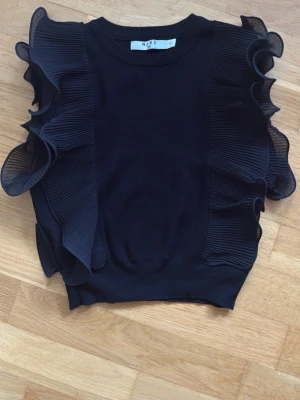 Svart topp med volang från NA-KD - Supersnygg svart topp från NA-KD med breda volangdetaljer längs axlarna. Toppen har rund halsringning och är tillverkad i ett mjukt material som känns bekvämt mot huden. Perfekt för dig som vill sticka ut lite extra.