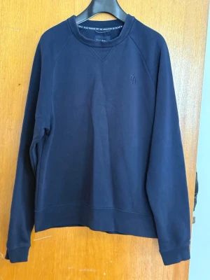 Mörkblå sweatshirt med RM-detalj - En stilren mörkblå sweatshirt med diskret RM-brodyr på bröstet. Tröjan har rund halsringning, långa ärmar och ribbade muddar vid ärmslut och nederkant. Perfekt för en avslappnad look och enkel att matcha med jeans eller joggers.