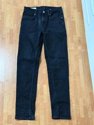 Svarta slim fit jeans - Snygga svarta slim fit jeans med klassisk femficksdesign och raka ben. Jeansen har bälteshällor, knappgylf och är tillverkade i ett mjukt denimtyg som sitter skönt. Perfekta för dig som gillar en stilren och enkel look.