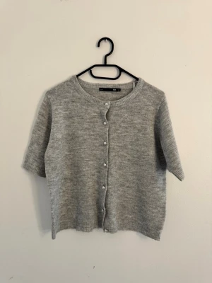 Ljusgrå kortärmad mysig tröja - Mysig ljusgrå tröja med korta ärmar och rund hals. Tröjan har knappar framtill och är stickad i ett mjukt material som ger en avslappnad look. Är lite varmare en än vanlig t-shirt.