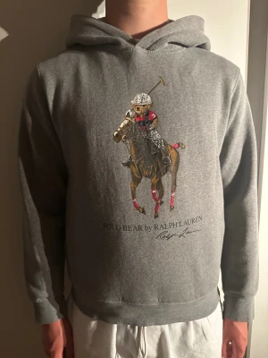 Grå hoodie Polo Bear Ralph Lauren - Snygg grå hoodie från Ralph Lauren med Polo Bear-tryck framtill där björnen rider häst i poloutstyrsel. Klassisk huva och ribbade muddar vid ärmar och nederkant. Perfekt för en avslappnad och trendig look.