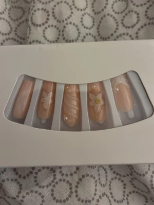 Nude press-on naglar med 3D-blommor - Säljer ett set press-on naglar i nude färg med 3D-blommor och gulddetaljer. Naglarna har en elegant, oval form och vissa är dekorerade med vita blommor och små guldfärgade pärlor. Materialet är plast och de är enkla att applicera själv.