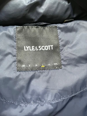 Svart dunväst från Lyle & Scott - Snygg svart dunväst från Lyle & Scott med hög krage och två stora fickor framtill. Västen har en quiltad design och det klassiska gula logomärket på bröstet. Perfekt för lager-på-lager och håller dig varm under kyliga dagar.