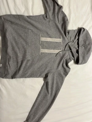 Grå hoodie med vita ränder - Snygg grå hoodie med två vita vertikala ränder på bröstet. Klassisk känguruficka framtill och justerbar huva med snören. Tillverkad i mjukt sweatshirtmaterial som är skönt mot huden. Perfekt för chill dagar eller när du vill ha en clean look.