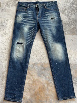 Dsquared2 jeans - Säljer ett par blå jeans med slitningar, hål på låret och coola färgstänk. Jeansen har en tvättad look med ljusa partier och raka ben. Perfekta för dig som gillar en avslappnad och trendig stil.