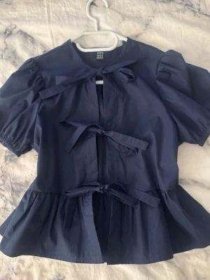Mörkblå blus med knytband - Snygg mörkblå blus med puffiga korta ärmar och tre dekorativa knytband framtill. Blusen har en lätt utsvängd midja och rund halsringning. Tillverkad i ett tunt, vävt material som ger en luftig känsla. Perfekt för en trendig och avslappnad look.